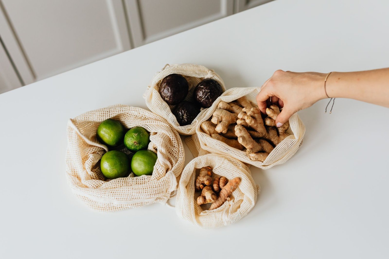 Mano che prende dello zenzero da un sacchetto della spesa riutilizzabile, con lime e avocado sul tavolo