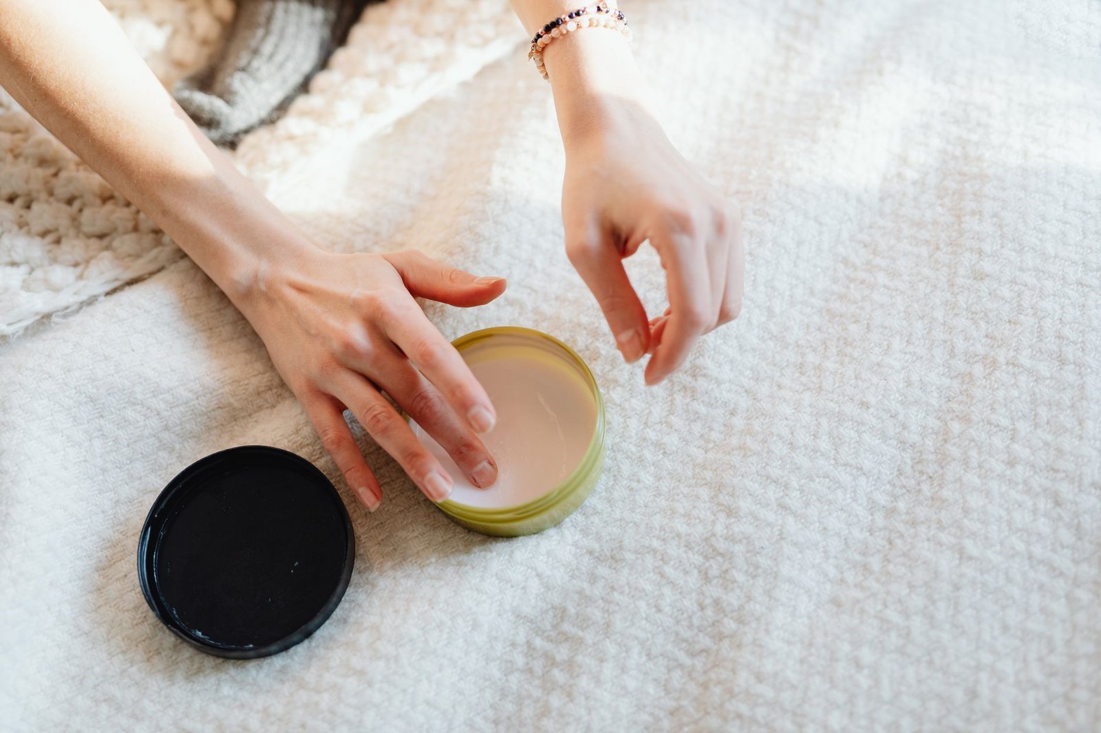 Mani che aprono un vasetto di crema su una coperta morbida di casa