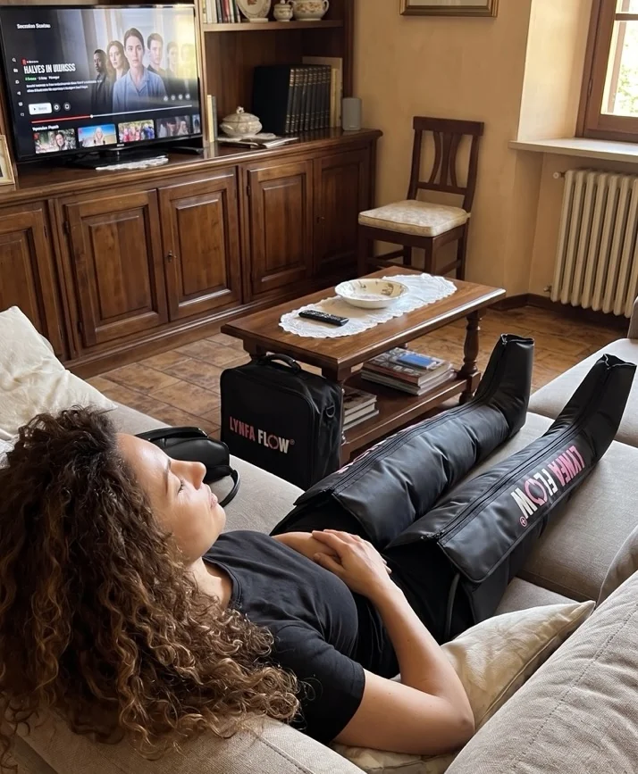 Donna rilassata sul divano con i gambali LynfaFlow e la borsa accanto, mentre guarda la TV
